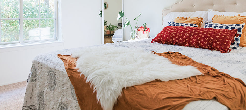 A room scene with a bed that is dressed in white with colorful throws and pillows.  The walls are white, and we can see plants both inside and outside the window.