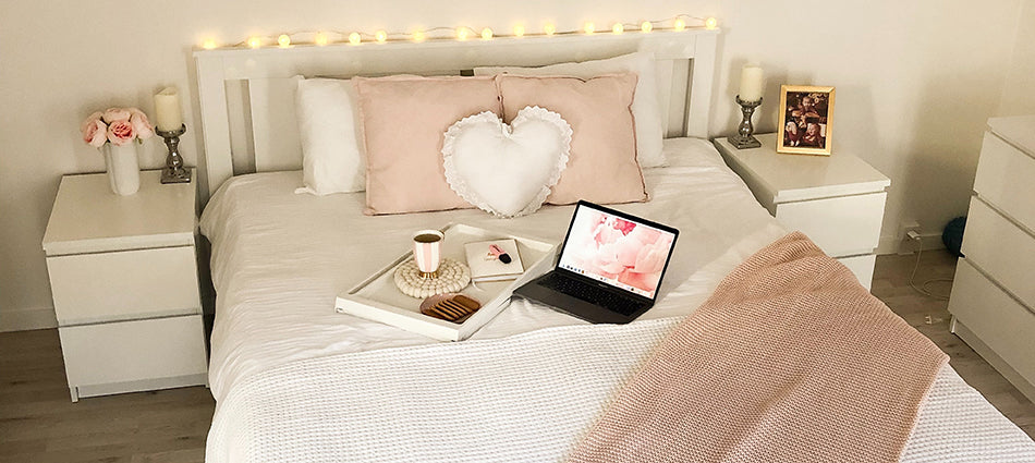 A room scene with a bed dressed in white with pink throws and pillows.  A heart-shaped accent pillow and string lights complete the look.  An open laptop and coffee tray suggest this is a dorm room.