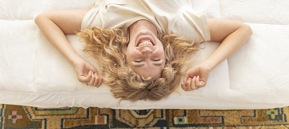 A woman is lying on a white mattress, shown from above. She is laughing joyfully.