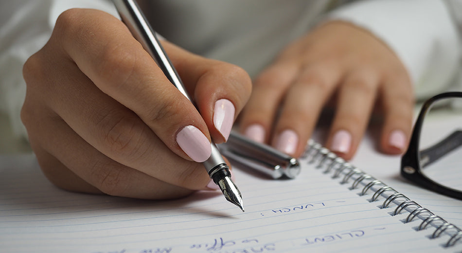 A close-up of a woman’s hand.  She has a pink manicure and is writing in a notebook with a fountain pen.  Her glasses sit on one half of the notebook.
