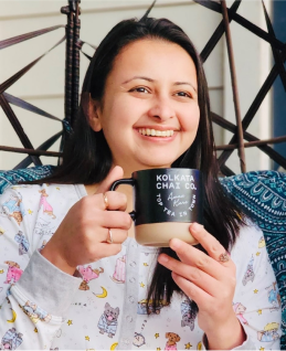 A woman smiling and holding a mug while sitting in a cozy setting.