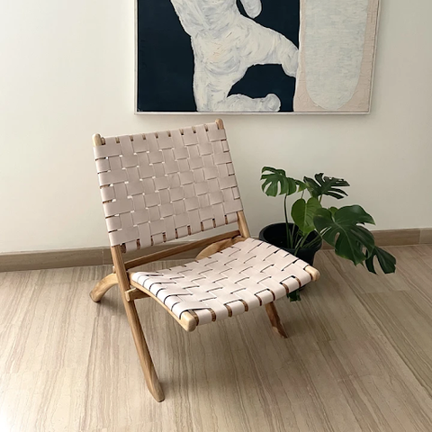 A beige foldable rattan chair placed in front of a painting and beside a plant