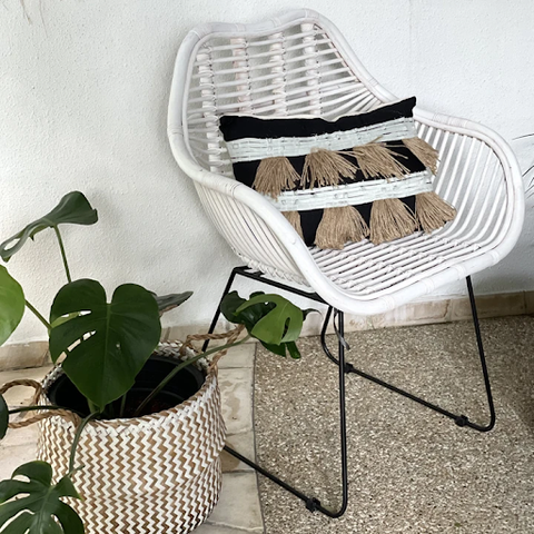 A white rattan chair with black legs coupled with a black pillow is placed beside a plant in a large rattan pot