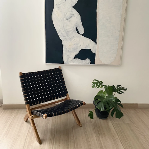 A black foldable rattan chair is placed beside a black pot plant and below a painting