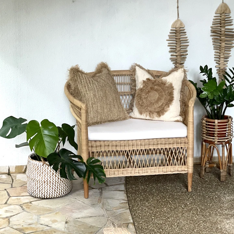 A two-seater rattan chair with two pillows and a broad white cushioned seat placed between two potted plants