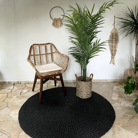 A natural rattan chair with a white cushioned seat placed nearly outside a black carpet and beside a plant in a rattan pot
