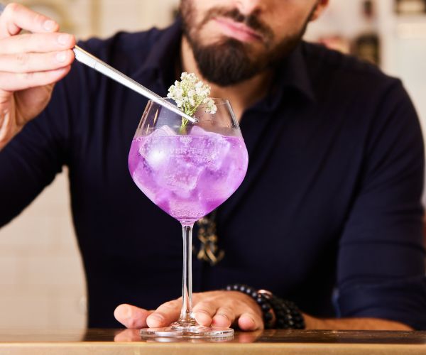 Bartender garnishing a purple cocktail with edible flower using a tweezer