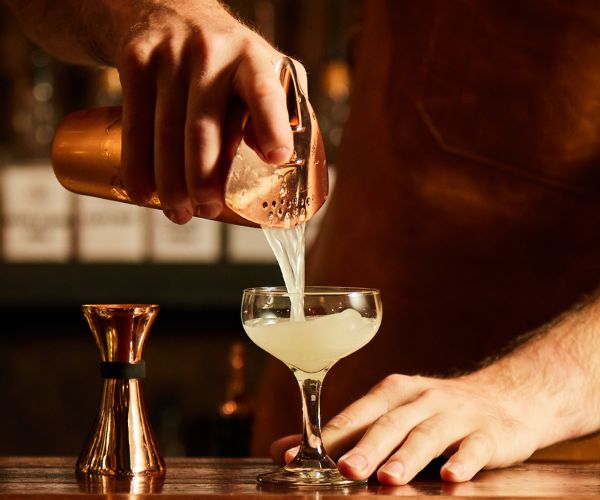 Bartender pouring shaken cocktail into a glass with jigger on the side