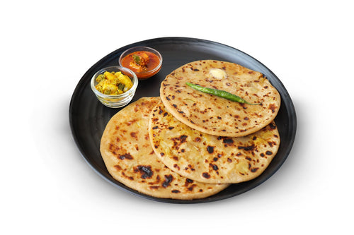 Three aloo paratha on a black plate with one green chilli and butter upon it served with some pickle and aloo vegetable mix.