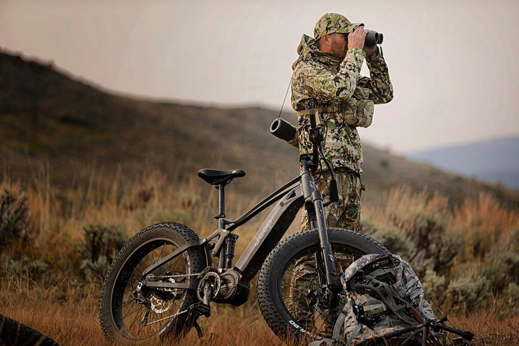 Un chasseur debout à côté de son vélo de chasse éclectique QuietKat Jeep cherche du gibier à travers une paire de jumelles