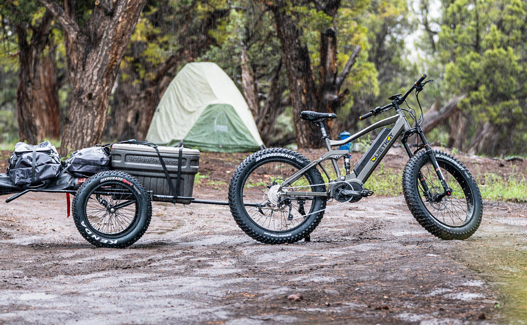 A QuietKat electric bike with a two wheel trailer is parked on a dirt trail with a tent in the background.