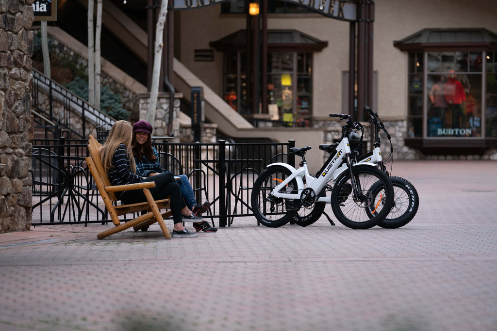 Two women sit on a bench by their parked QuietKat electric bikes near a cafe.
