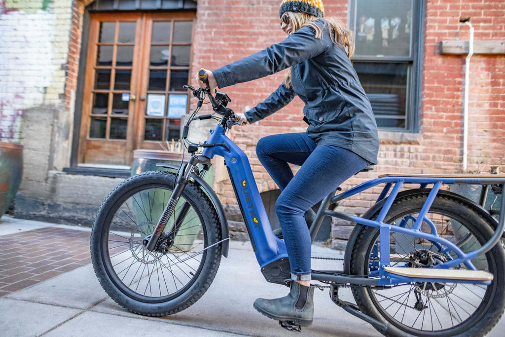 Une femme chevauche son vélo cargo bleu QuietKat Sherpa dans une rue de la ville.