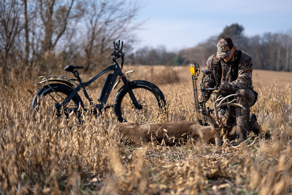 Un chasseur à l'arc s'agenouille à côté d'un mâle tombé avec un vélo de chasse électrique QuietKat en arrière-plan