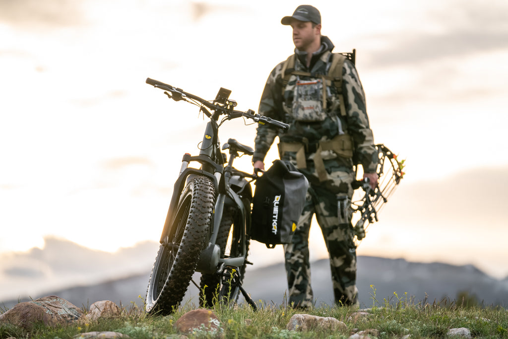 A bow hunter looks out ove rteh landscape while standing by his QuietKat electric bike with pannier bags.