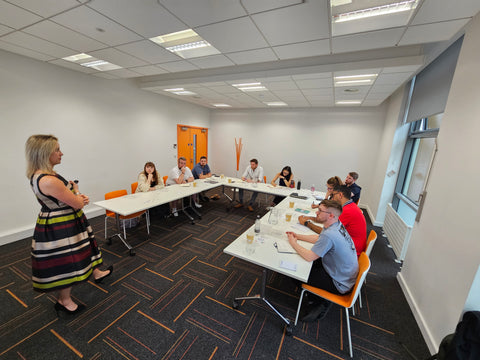 Team training - this photo shows a group of learners from a business in a classroom setting around white tables looking at Jessica Williams leading the training.