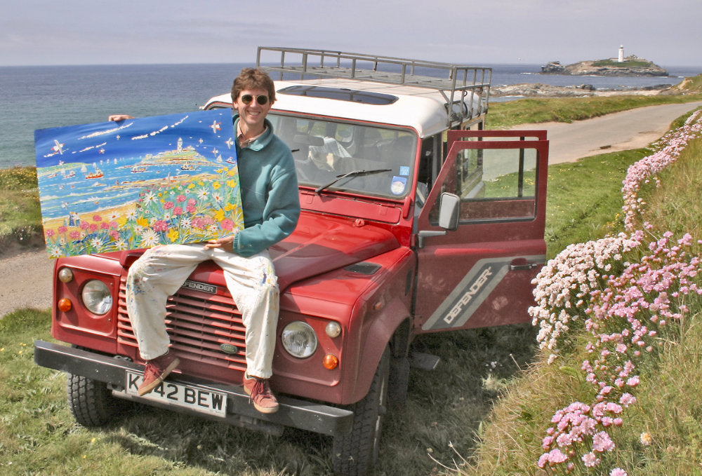 Artist John Dyer sitting on his Landrover Defender, holding his painting of Godrevy