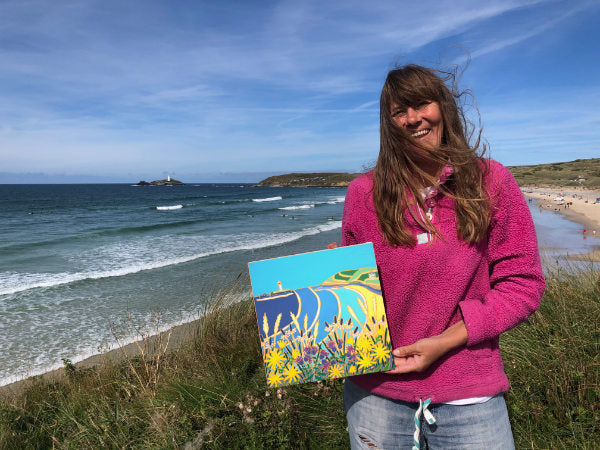 Cornish artist Joanne short standing on the cliffs at Gwithian in Cornwall holding her painting of Godevy lighthouse