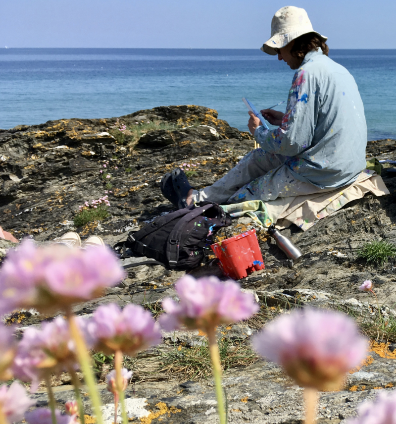 Artist John Dyer at work sitting on the rocks and painting the Roseland in Cornwall. Sea pinks in the foreground