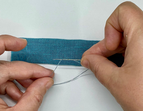 Two hands, pulling a threaded needle through the last loop on a thread chain, securing the chain to a blue dress strap with a knot. 