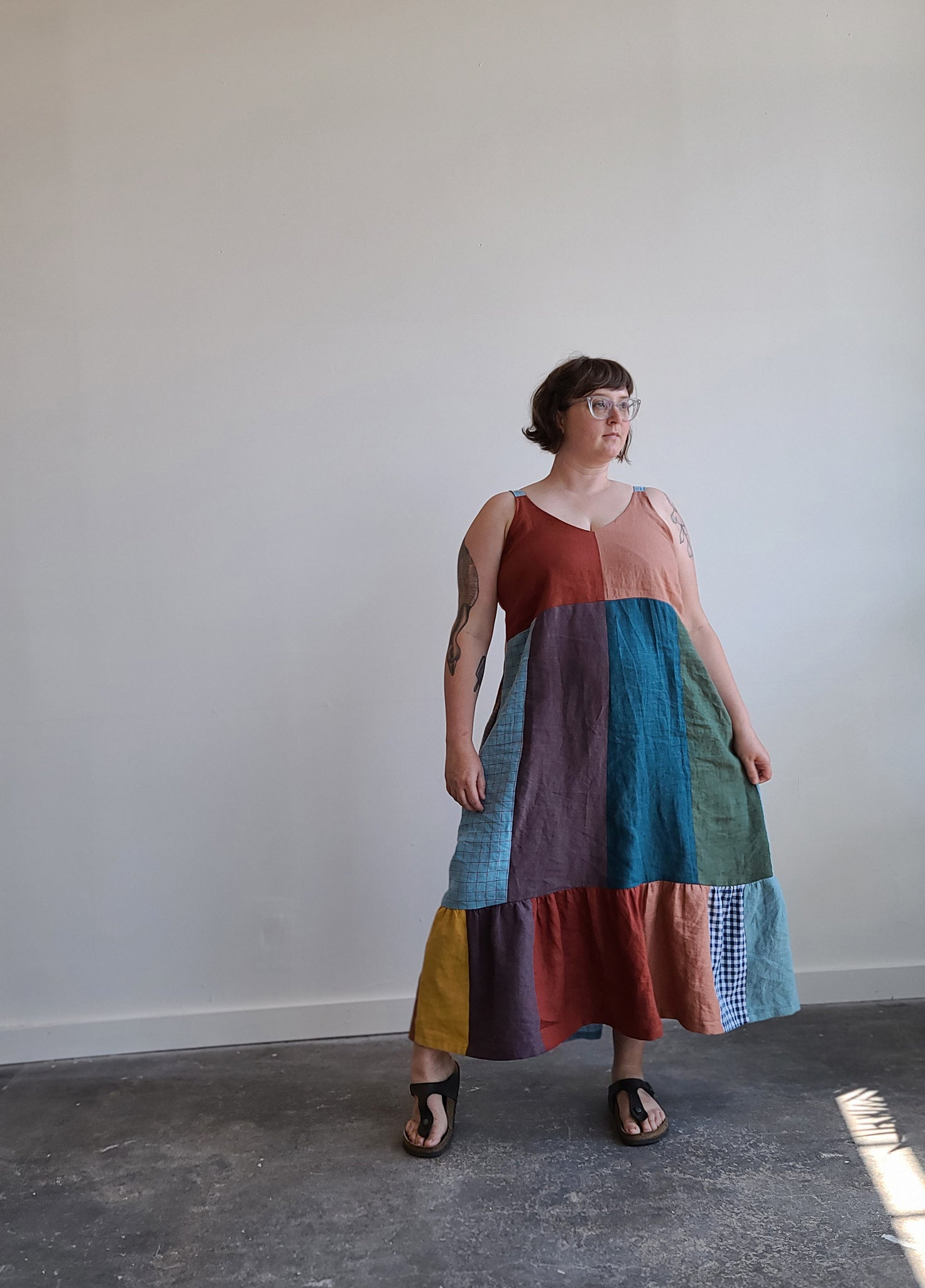 Windsor, a midsize white woman, standing in front a white wall, wearing a multicolored linen patchwork Sauvie Sundress