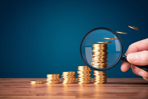 A hand holds a magnifying glass against a stack of coins on a table.