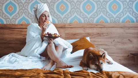 A girl with a towel on her head and a dog on the bed eats fruit for breakfast.