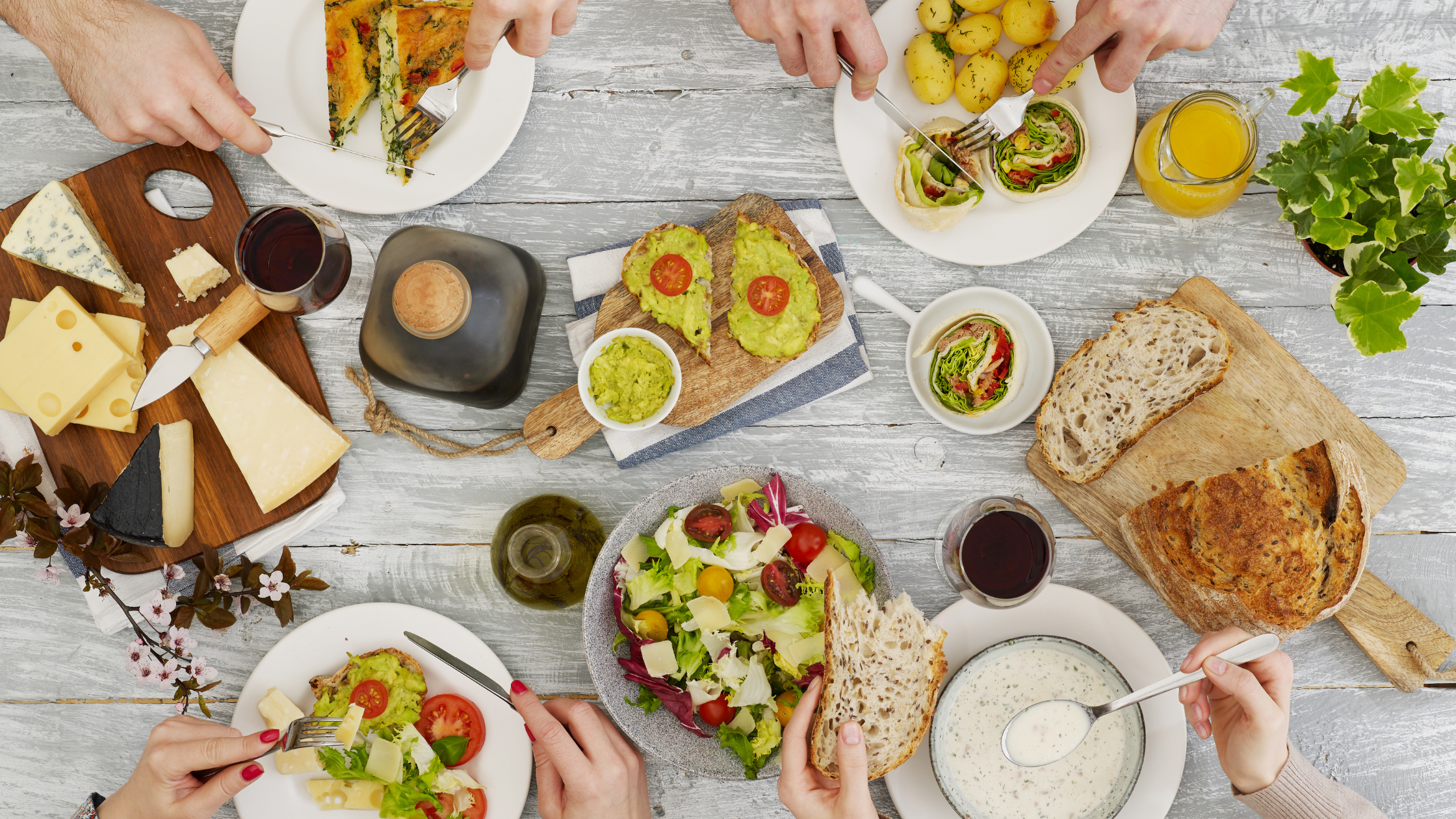 A table and people's hands eating food and drinking wine, the table has a cheese board, bread, salads, olive oils