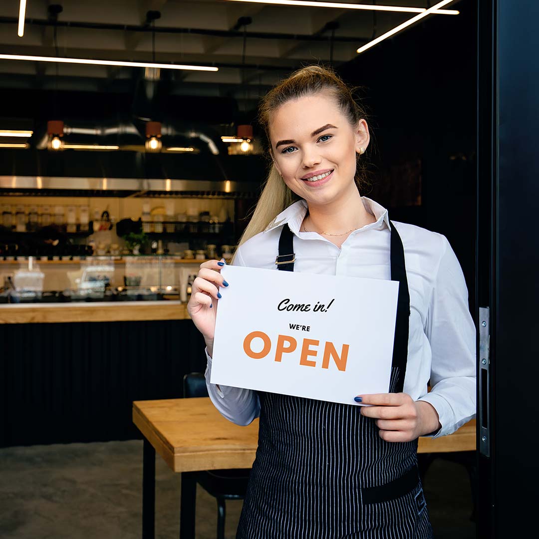An image of a woman holding an “open” sign in front of a restaurant.