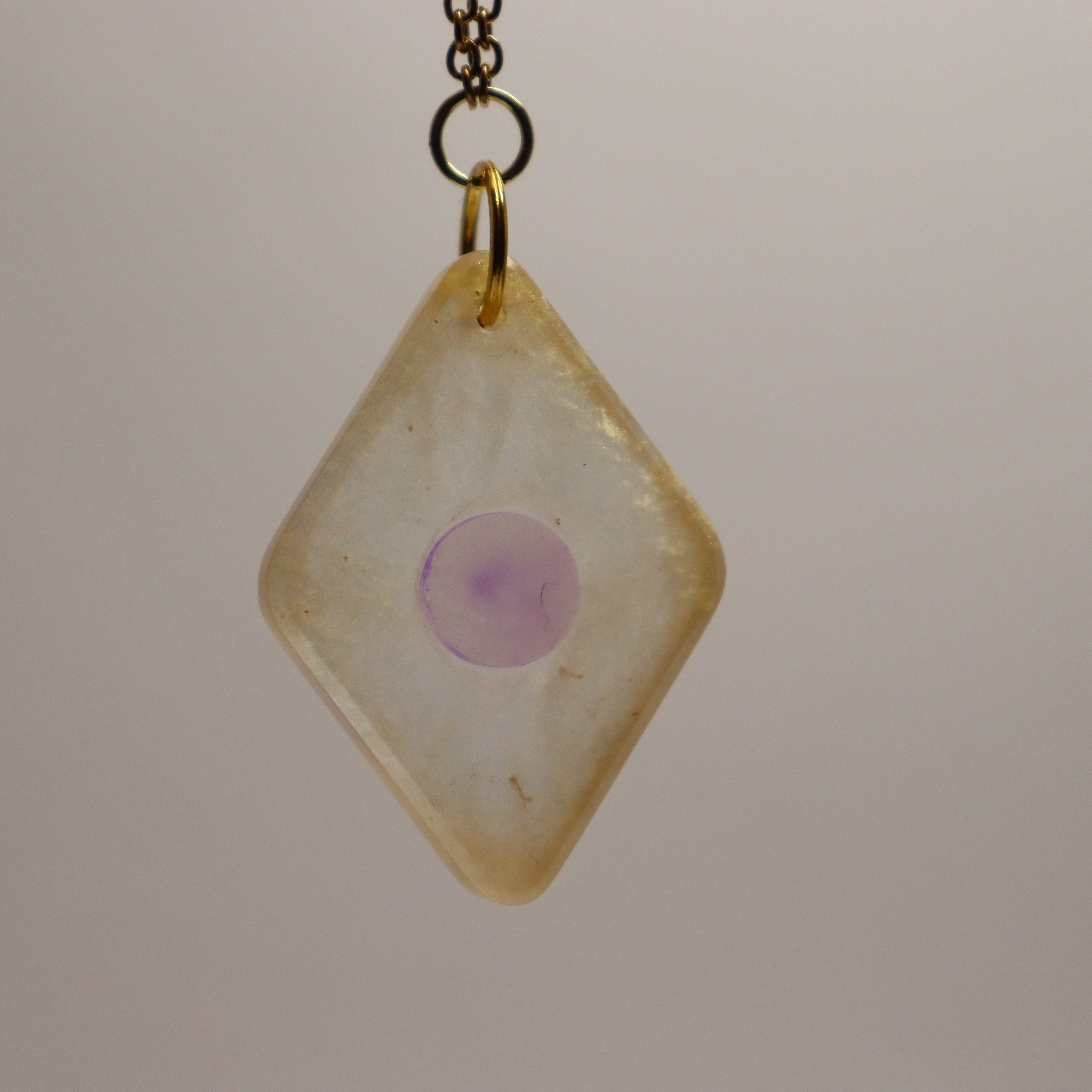 A diamond shaped necklace hangs on a gold chain. The necklace has a gold exterior that becomes more transparent closer to the centre and a light purple circle in the middle.
