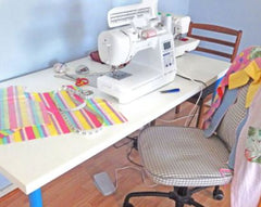A shot of a sewing table with a multicoloured fabric project. There are sewing machines and tools on the table. There are chairs around the table.