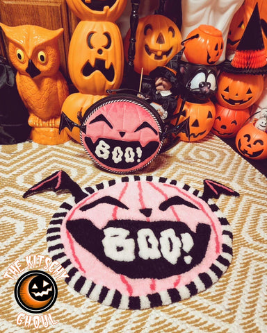 pink pumpkin rug with boo text in mouth beside a matching purse, a background of orange pumpkin decorations