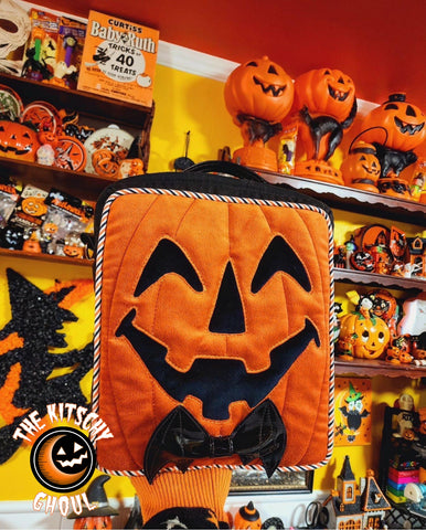 A square pumpkin purse with a bat bow tie being held up against a background of halloween decorations