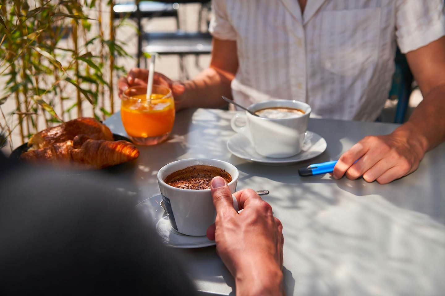 Two people sitting at an outdoor cafe table drinking coffee, one with a vape in hand
