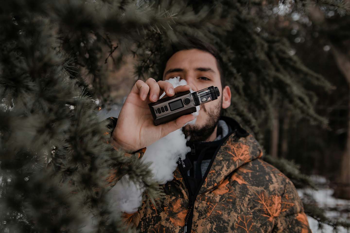 man vaping in brown and black camouflage jacket holding black and silver vape