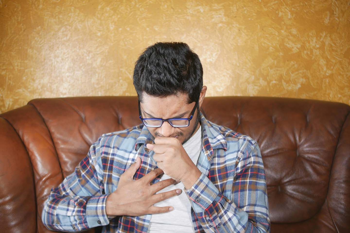 A man sitting on brown leather sofa and holding his chest while coughing