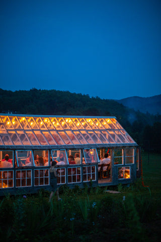 The glasshouse at night, illuminated from within against a blue-green, hilly backdrop. 