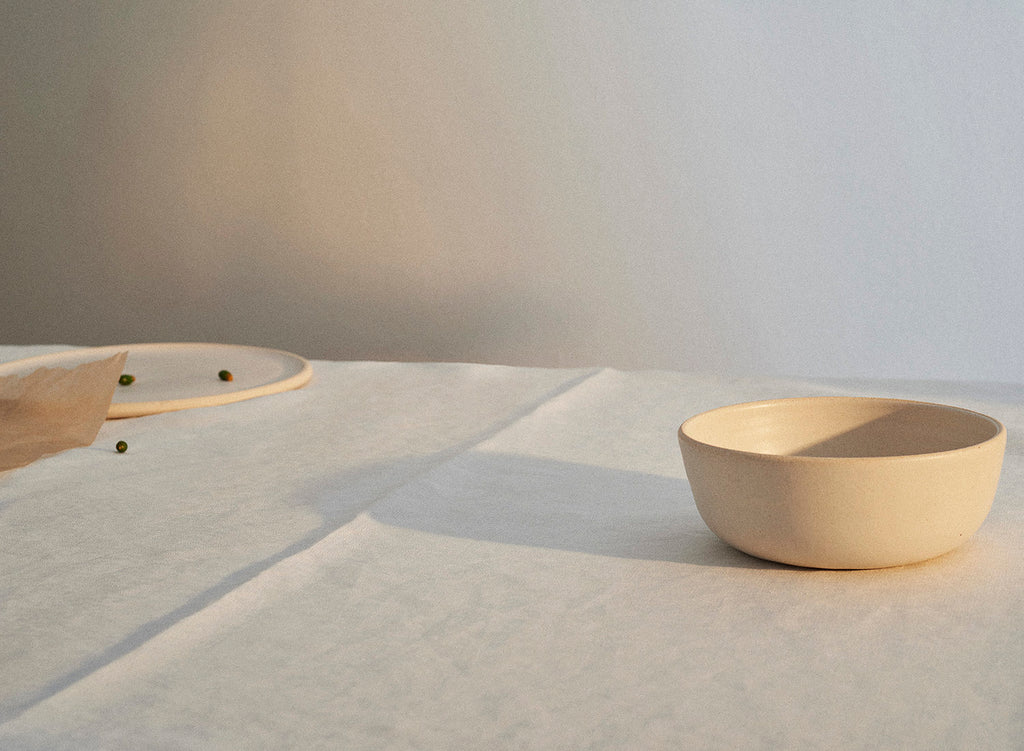A handmade Sheldon Ceramics Silverlake Low Profile Bowl in Eggshell casts a long shadow across a white linen tablecloth. 