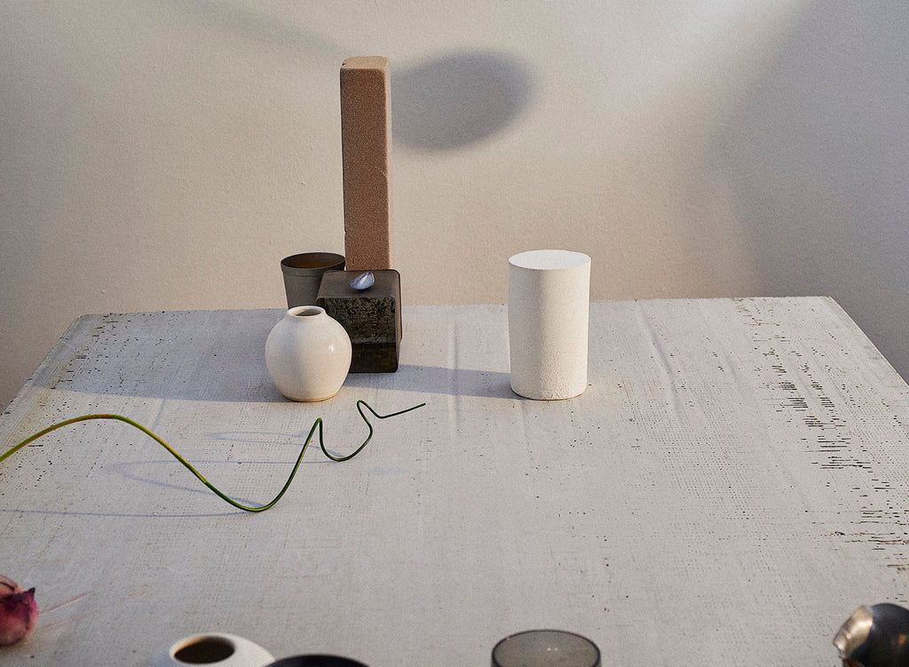 A white table scattered with an assortment of objects: a handmade ceramic vase, a brick, a length of green wire, and an aluminum tin.