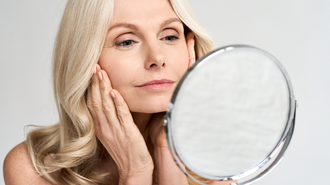 A blonde older woman looks at her skin in the mirror, it is bright and clear.