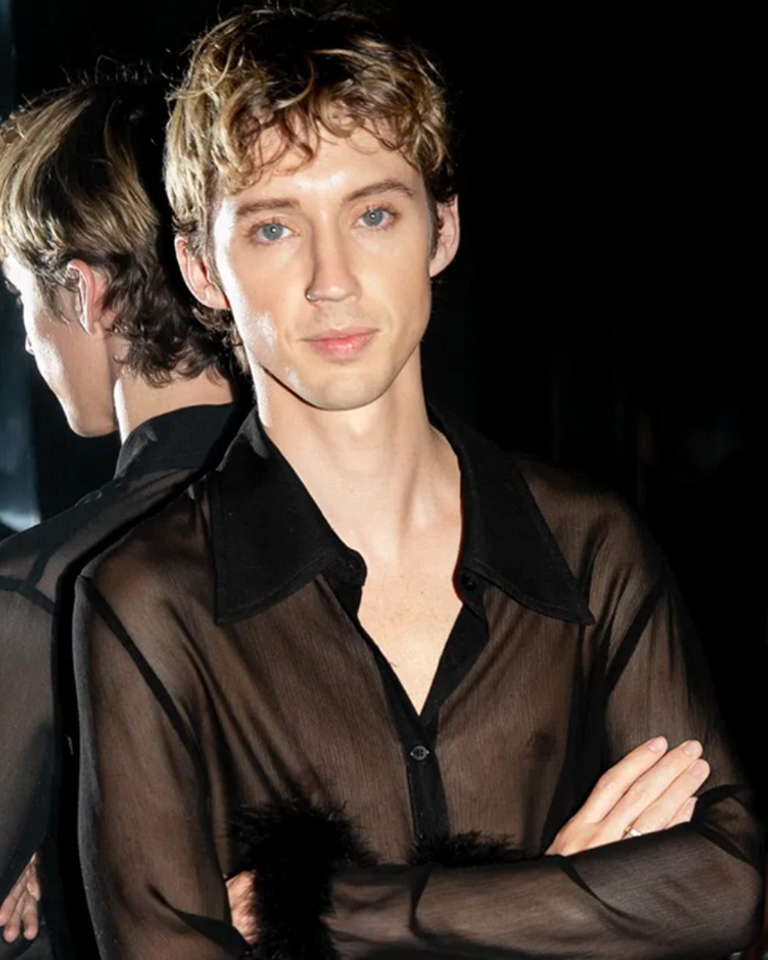 Person with short curly hair, wearing a sheer black shirt, stands with folded arms in front of a mirror.