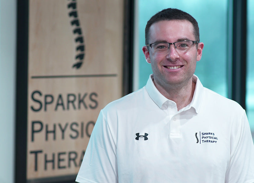 A man in a white Sparks Physical Therapy shirt standing indoors by the clinic sign.