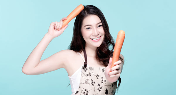 woman rubbing carrot on her hair and holding another carrot standing in front of a blue background