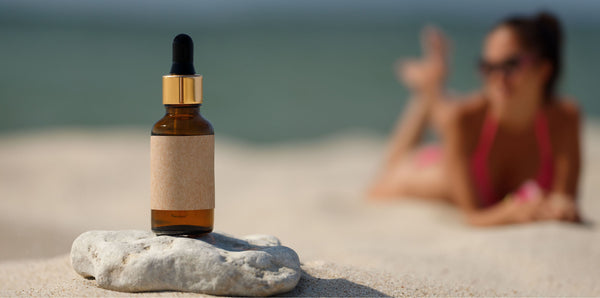 essential oil bottle on rock on sandy beach with woman in bathing suit in the background