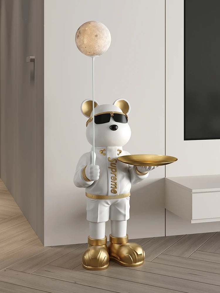 A decorative sculpture of a white bear in sunglasses, wearing a white shirt, golden shoes, and holding a tray beside a balloon shaped like the moon that emits a soothing glow.