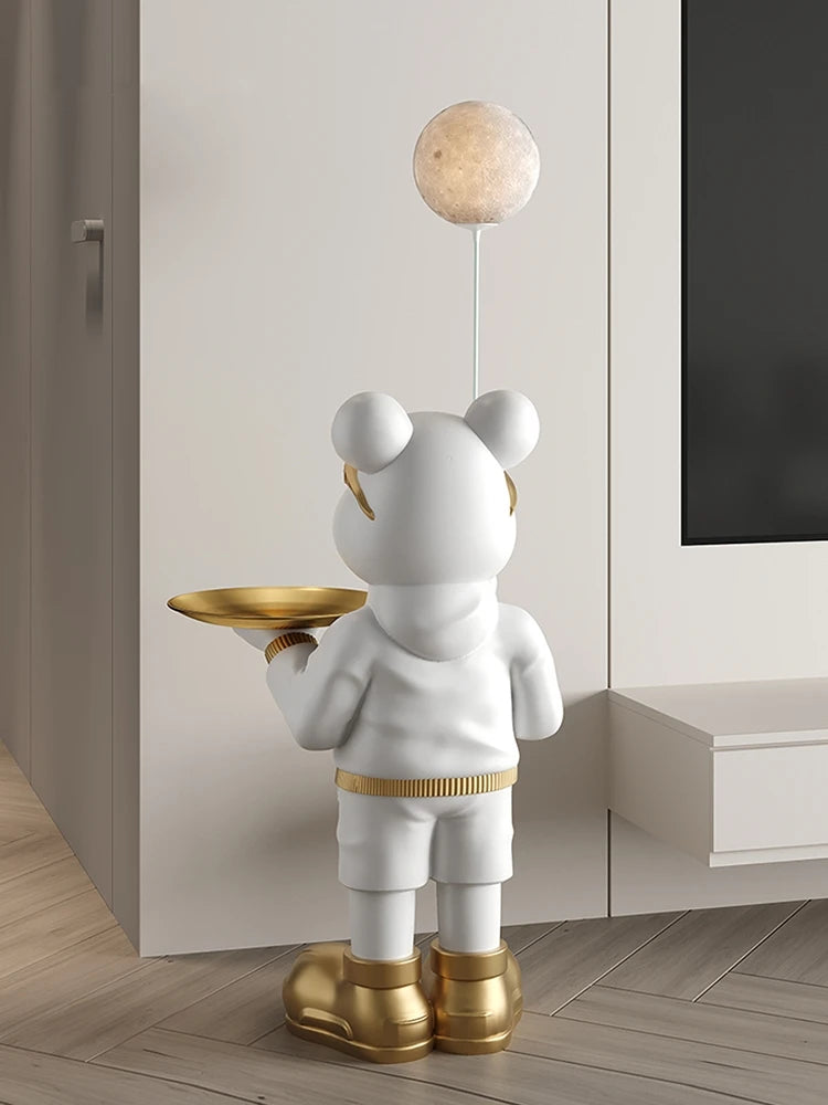 Decorative sculpture of a cartoon bear in a white outfit and gold shoes, holding a tray, with a soothing glow moon lamp above its head in a modern room.