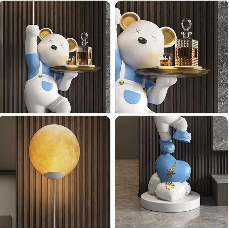 Decorative display featuring an adorable bear statue in a waiter outfit, holding a tray with gold trophies, set against a textured wall with a spherical lamp nearby.