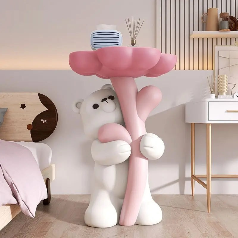 A cartoonish bear sculpture holding a large pink floral tabletop with decor items on top in a stylish room.