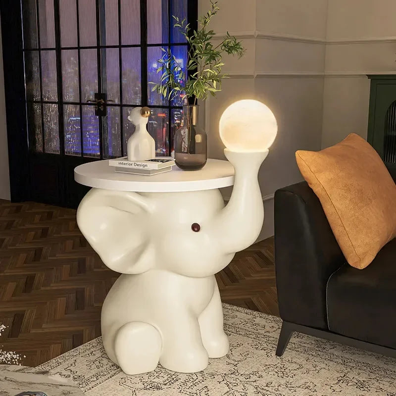 An elephant-shaped bedside table lamp with a round light as the trunk, supporting a small tabletop with decorative items, in a bedroom with a sofa and herringbone floor.