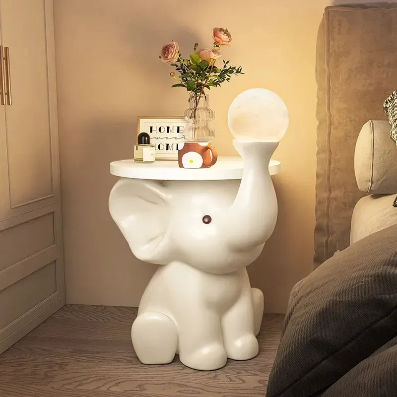 A white elephant-shaped bedside table holding a vase with flowers, a book, and small objects, illuminated by a warm light in the bedroom.